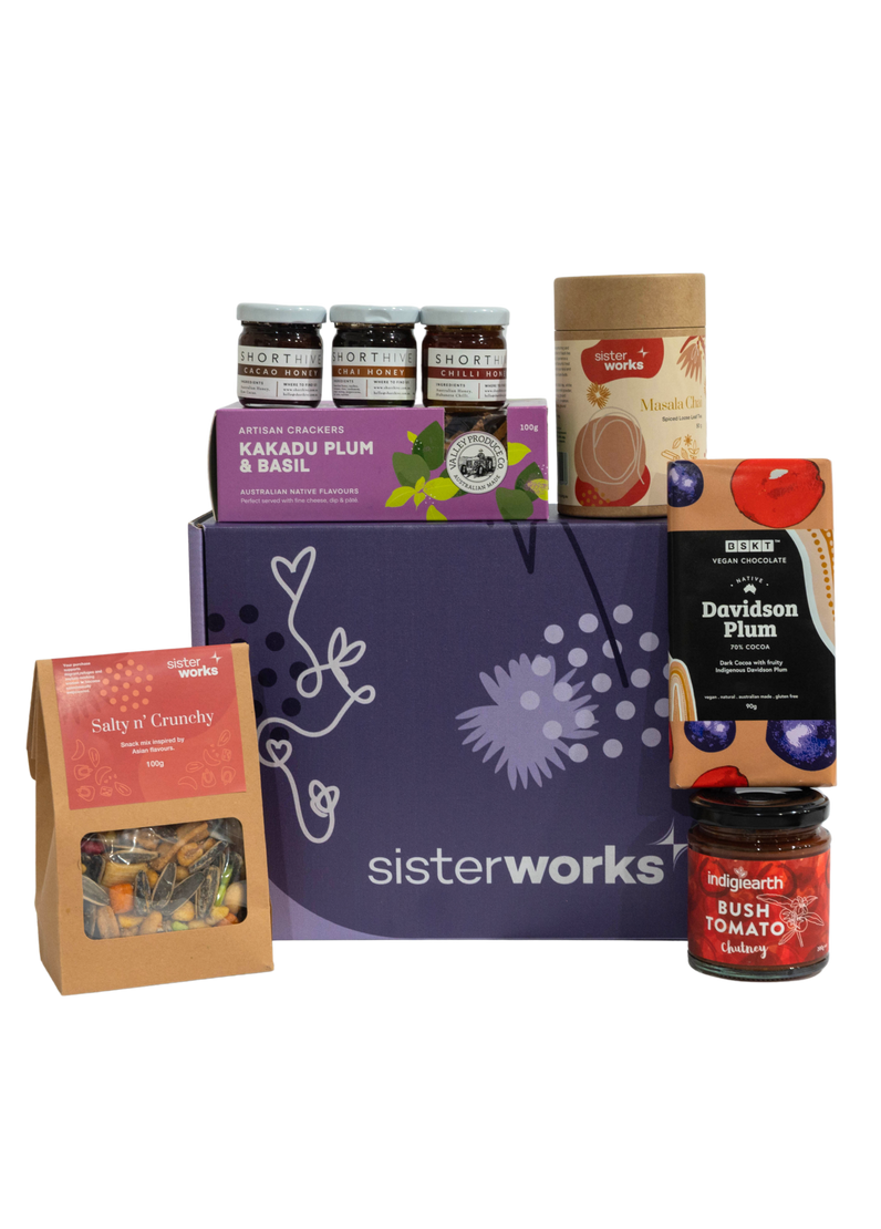 An overhead view of the open SisterWorks Foodie Hamper. The contents are neatly arranged in a box, including a jar of Eggplant Pickle, a 3-pack of ShortHive Infused Honey, a pouch of Cinnamon Myrtle Black Tea, a bag of Salty n' Crunchy Mix, and a box of Kakadu Plum Co. Crackers. The collection highlights a perfect gourmet gift set for a foodie