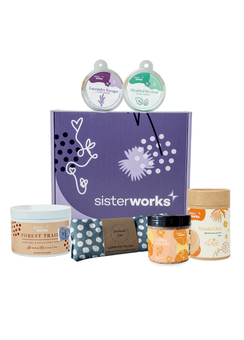 An overhead view of the open SisterWorks Aromatherapy Bliss Hamper. The contents are neatly arranged inside a box, including a pouch of Masala Chai, a Lavender Escape Shower Steamer, a Menthol Revival Shower Steamer, a Forest Trails Candle, an eye pillow, and a jar of uplifting bath salts, highlighting an ethical gift set for relaxation and self-care.