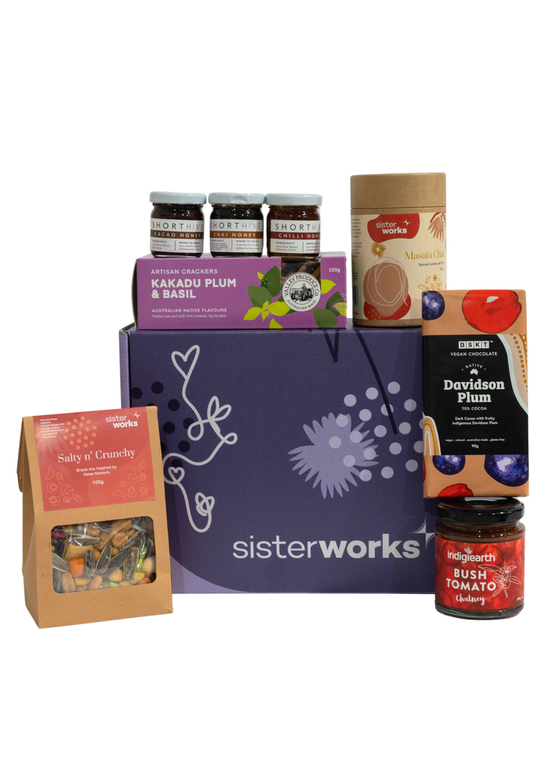 A SisterWorks Aussie Holiday Hamper. The box contains STREAT Gingerbread Stars, SisterWorks Raspberry Jam, BSKT Davidson Plum Vegan Chocolate, SisterWorks Lemon Myrtle Green Tea, and Australian Icon Coasters. The items are arranged neatly in a box, highlighting a festive and ethical gift set.