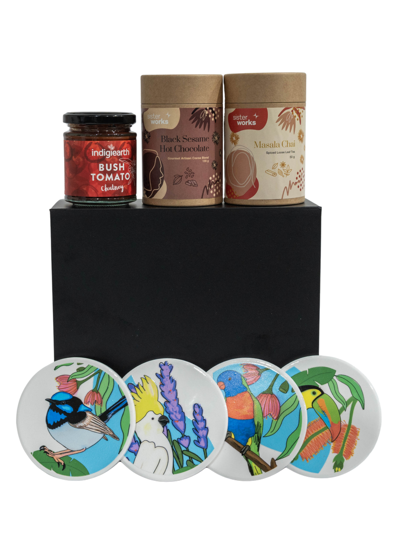 Front view of the SisterWorks Cultural Fusion Hamper contents laid out. Features SisterWorks Black Sesame Hot Chocolate, Masala Chai, bush tomato chutney,  and four vibrant Cultural Fusion Coasters by artist Marynel Jaimes. The sleek black premium gift box is visible, signifying a luxurious, ethical corporate gift or a unique present celebrating global artistry.