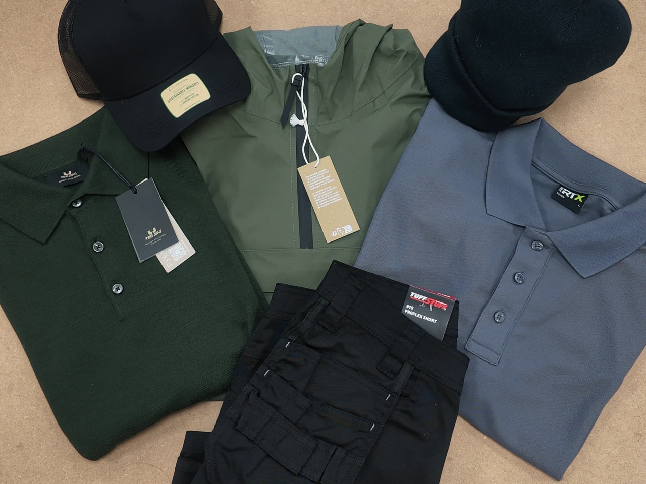 A flat-lay of workwear laid out together — a knitted polo, a waterproof jacket, work trousers, a performance polo and two caps