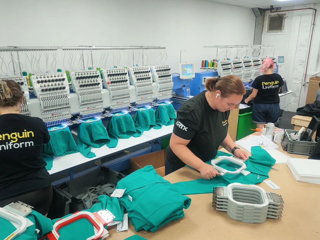 The Penguin Uniform embroidery floor in use — a line of multi-head machines running green polo shirts while three members of the team hoop garments and prepare the next run