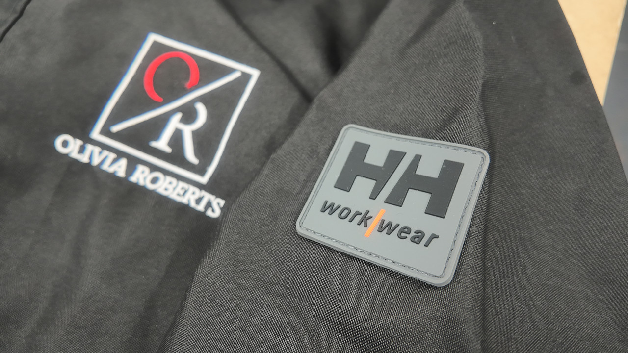 A customer logo applied to the chest of a black technical waterproof jacket, beside the garment manufacturer's badge