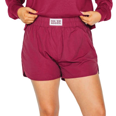 Scarlet & Gold maroon nylon shorts featuring an elastic waistband, curved side hems, relaxed fit, and a small white “Gig ’Em Aggies” label centered along the front waistband.