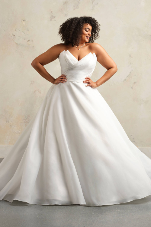 Plus-size bride wearing the Maggie Sottero Tinsley ball gown, showcasing the full organza skirt and pointed neckline. Plus-size bride wearing the Maggie Sottero Tinsley ball gown, showcasing the full organza skirt and pointed neckline.