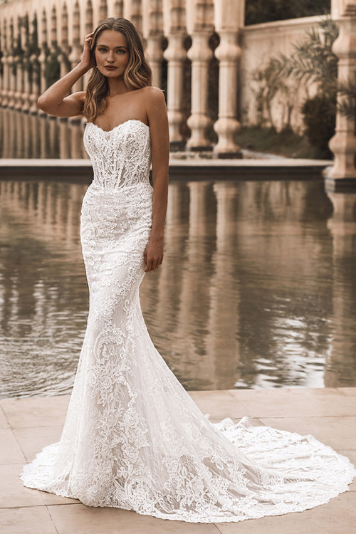 Bride wearing Enzoani Vail fitted gown with lace detailing, front view by water. Bride wearing Enzoani Vail fitted gown with lace detailing, front view by water.