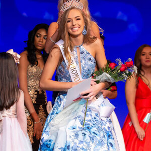 Miss pageant winner in blue and white floral gown with crown and flowers