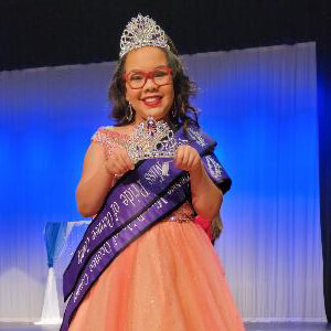 Junior pageant winner in peach gown wearing glasses and crown