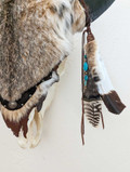 Closeup section of Bison skull wall hanging covered in bobcat fur showing detail of fur, feathers, leather tassels, and strands with turquoise and small decorative beads.