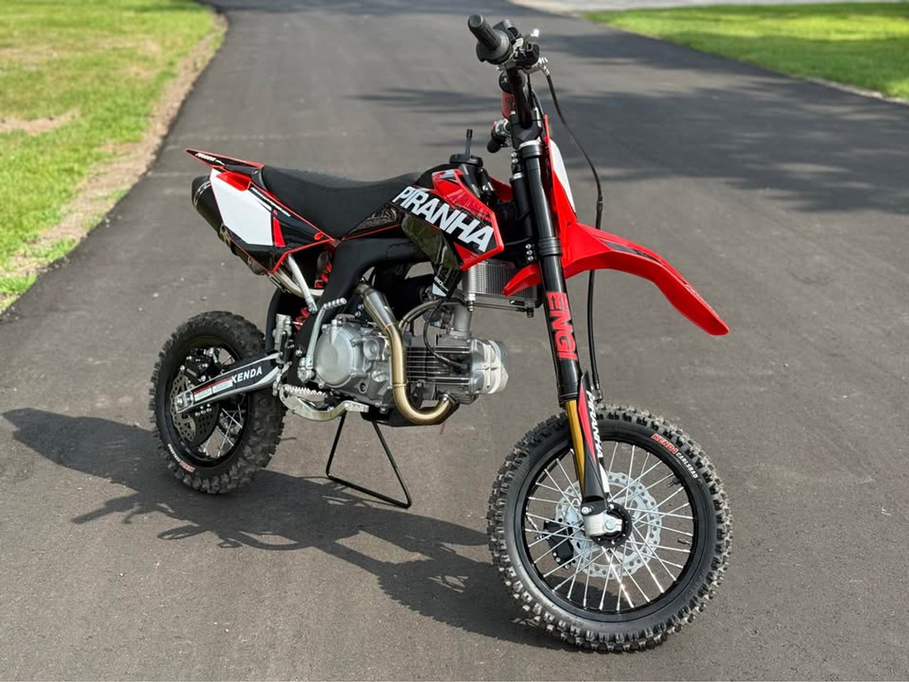 2024 Piranha Daytona 190-4V DE (Dual Exhaust), a high-performance competition pit bike designed for serious riders. The bike features a sleek and aggressive design with a dual exhaust system, emphasizing its power and performance. The Daytona Anima 190 4-valve engine and oversized oil cooler are highlighted, showcasing the bike’s robust power and efficient cooling. The ENGI adjustable suspension (800mm front forks and 330mm rear shock) is prominently displayed, underscoring the bike’s superior handling and shock absorption. The 14-inch front and 12-inch rear aluminum rims with Kenda Carlsbad tires add to the bike’s durability and traction. Overall, the image captures the essence of a high-performance, competition-ready pit bike built to dominate the track.