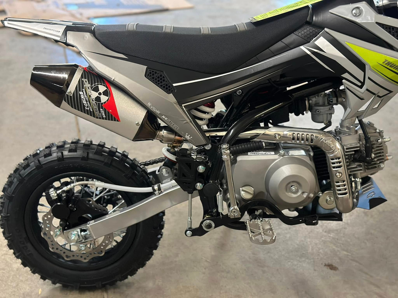 Thumpstar TSK 90cc mini bike, featuring its sleek mini stealth plastics, compact frame, and bold design. The bike is displayed in an action-ready stance, highlighting its inverted front suspension, oversized disc brakes, and Pipe Bomb muffler. The wrap-around frame and waterproof wiring are subtly visible, emphasizing the bike’s durability. The image also highlights the adjustable throttle limiter and emergency lanyard kill switch, key safety features for junior riders.