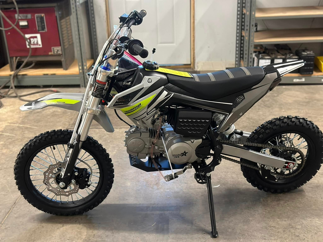 Thumpstar TSC 125 pit bike, highlighting its signature stealth plastics, wrap-around frame, and gripper striped seat. The bike is displayed in an action-ready stance, emphasizing its conventional front forks, rear mono shock, and front and rear disc brakes. The forged triple clamps and stator guard are subtly visible, underscoring the bike’s durability and safety features. The Mikuni VM22 carburetor and standard exhaust system are also highlighted, showcasing the bike’s performance enhancements.