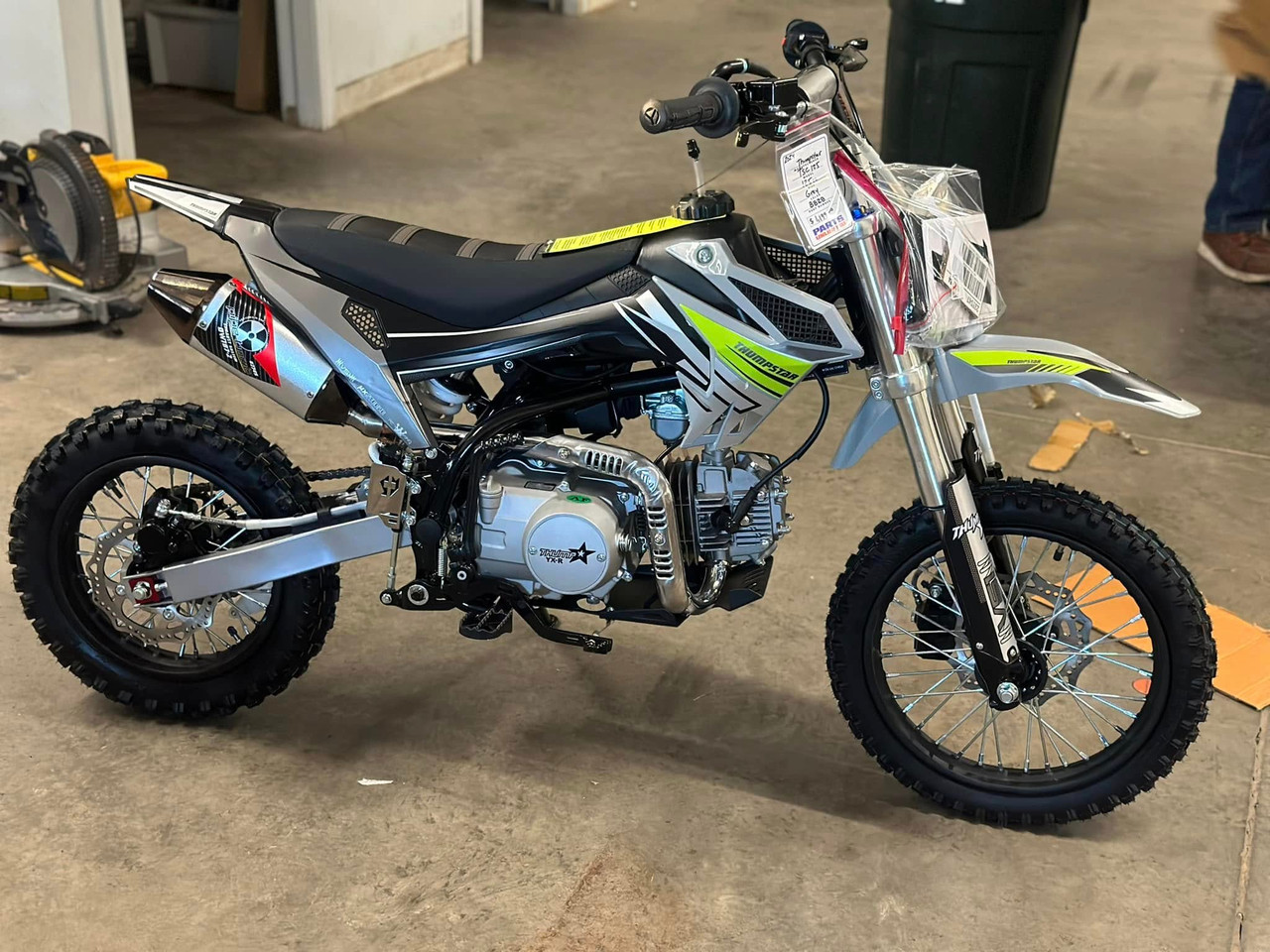 Thumpstar TSC 125 pit bike, highlighting its signature stealth plastics, wrap-around frame, and gripper striped seat. The bike is displayed in an action-ready stance, emphasizing its conventional front forks, rear mono shock, and front and rear disc brakes. The forged triple clamps and stator guard are subtly visible, underscoring the bike’s durability and safety features. The Mikuni VM22 carburetor and standard exhaust system are also highlighted, showcasing the bike’s performance enhancements.