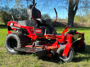 2025 Toro 60" TITAN MyRIDE Zero Turn Mower (76614) showcasing its rugged, commercial-grade design and premium features. The mower features a heavy-duty IronForged® 10-gauge steel cutting deck, built to withstand tough mowing conditions with reinforced belts and massive spindles. The commercial-style I-beam front end highlights its extreme durability and strength, while the 22" rear tires with aggressive treads ensure maximum traction on any terrain. The MyRIDE® Suspension System is prominently displayed, isolated from the frame to provide a smooth, comfortable ride. The treaded 13" front casters add stability and a rugged aesthetic, and the 20" gaming-style seat offers exceptional comfort for long mowing sessions. The rubber discharge chute is visible, designed to flex and protect landscaping while delivering a clean cut. This image captures the Toro TITAN's powerful Kohler KT745 26 HP (747cc) engine, pro-level drop pin height adjustment system, and overall dominance as a top-tier residential zero-turn mower. Perfect for homeowners seeking commercial-grade performance and comfort in a mower.