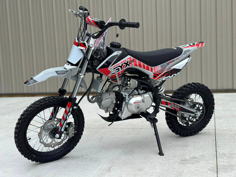 Syx Moto PAD125-1S 125cc semi-automatic pit bike featuring a black steel frame, aggressive new plastics design, and knobby off-road tires. The bike is shown in an off-road setting, highlighting its front hydraulic disc brake, remote oil cooler, and dual start system. The 14-inch front wheel and 12-inch rear wheel with heavy-duty spokes are clearly visible.