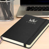 A black Moleskine Classic Hard Cover Notebook with a logo, placed on a desk beside a laptop and pen holder.