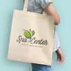 Natural beige tote bag featuring a logo with green leaf elements, held by a person against a light blue background.