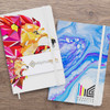 Two notebooks displayed side by side, one with a colourful lion design and the other in blue marbled pattern, both featuring logos.