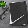 A black Moleskine Classic Hard Cover Notebook with a pen and glasses, placed on a textured surface with a plant.