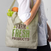 A cotton shoulder tote bag in light beige, featuring a logo and filled with lettuce and a drink bottle.