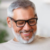 Malibu Blue Light Glasses in black, worn by a smiling man with grey hair and beard, indoors. The glasses have a logo.