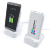 A white wireless power bank and charging station with a smartphone on a stand, displaying a charging battery icon.