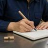 A cardboard pen in a natural finish, resting on a notebook beside a person writing with it.