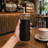 A glass tumbler with a black silicone sleeve, wooden lid, and straw, set on a wooden table with café surroundings.