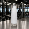 A drink bottle in sleek white with a stainless steel bottom, standing on a gym table. The bottle features a logo.