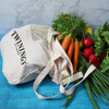 A cotton shopping tote in cream with a mesh top, filled with carrots, radishes, and fresh herbs.