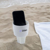 A white sand spike product holding a smartphone, placed on sand near a beach towel. The product features a branded logo.