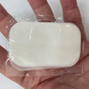 A hand holding a clear packaging containing a white, oval-shaped product. The product has a soft texture.
