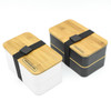 A two-layer lunch box set featuring a bamboo lid, one in white and one in black, secured with a black strap.