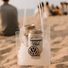 A clear cooler bag filled with ice and two cans, featuring a logo. Set on a sandy beach with people in the background.