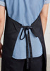 A bib apron in black, featuring a tie at the back and worn over a light blue shirt.