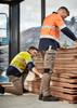 Two men in workwear, one in a yellow and navy shirt and the other in an orange and navy shirt, examine wooden panels while wearing beige stretch pants.