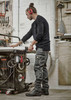 A worker in black Mens Streetworx Heritage Pants, operating machinery in a workshop, with tools and equipment in the background.