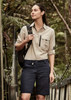A woman in a light beige shirt and navy shorts stands outdoors, with a backpack over her shoulder. The shorts feature a logo.