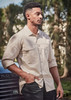 A lightweight long sleeve shirt in beige, featuring a front pocket and a logo. Set outdoors with greenery in the background.