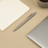 A stainless steel ballpoint pen rests beside a blank notepad and a laptop, with a cup of coffee in the background.