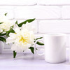 A white ceramic mug beside a peony flower on a light purple surface against a white brick background.