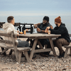 A cooler bag in black and white, sitting on a picnic table, with three people enjoying drinks and snacks.