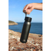 A drink bottle in matte black with a lid and attached strap, set against a coastal backdrop.
