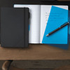 A black notebook with a blue folder and a pen sits on a wooden desk, featuring an open page with handwritten notes.