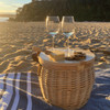 A wicker cooler basket in natural colour sits on a beach, with two wine glasses and a cheese board on top.