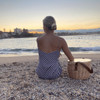 A wicker cooler basket in a light colour, positioned near a person on a beach at sunset. The scene features water and a cityscape.