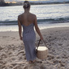 A wicker cooler basket with a wooden lid, carried by a person in a striped dress, on a beach at sunset.