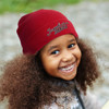 A red beanie with a logo, worn by a smiling child with curly hair, set against a natural background.