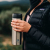 A stainless steel travel mug held by a person in a black puffer jacket, featuring a logo on its body.