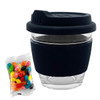 A glass coffee cup with a black silicone sleeve, accompanied by a packet of colourful jelly beans.